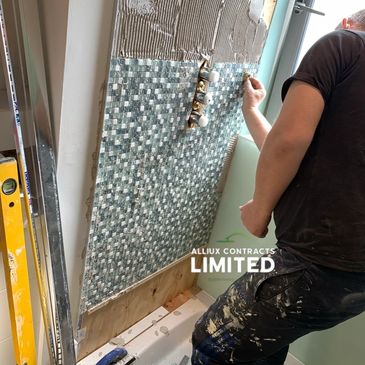 Worker installing mosaic tiles on a shower wall with tools nearby. Alliux Contracts Limited