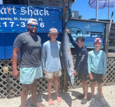 Four people proudly pose with a large caught fish at a fishing shack.