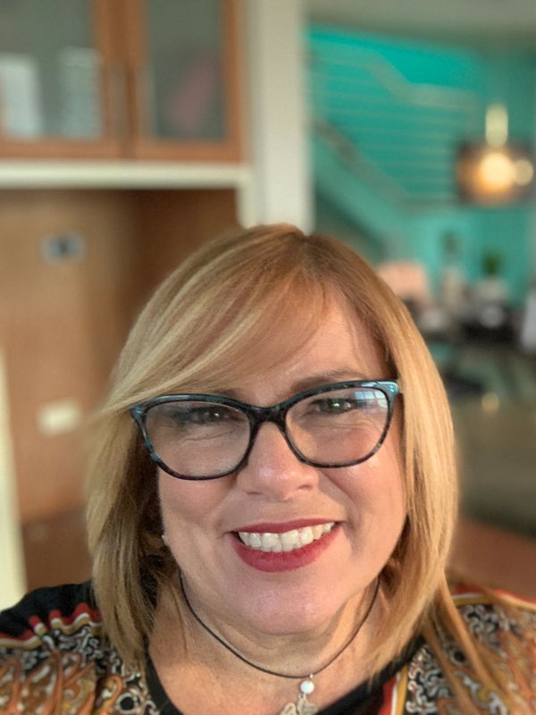 Smiling woman with glasses in a cozy, colorful living room with sunflowers in the background.
