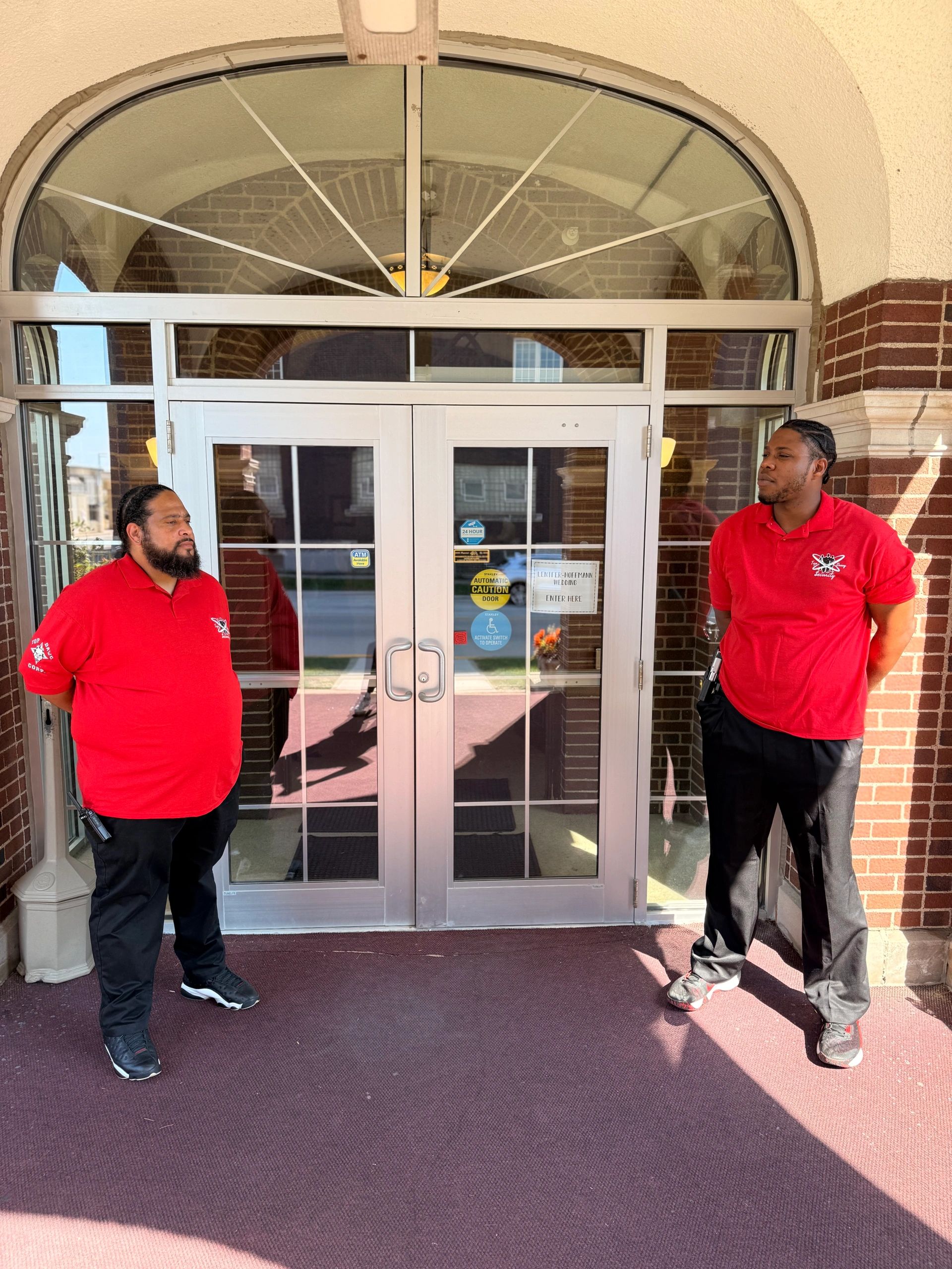 Two men in red shirts standing guard outside glass doors in a brick building.