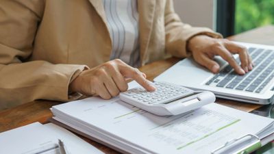 Person calculating finances with a calculator and laptop at a desk.