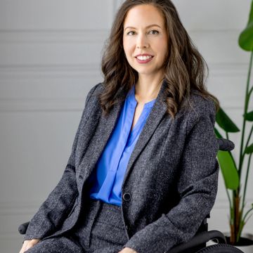 A photo of Dr. Sharon Surita wearing a gray suit, sitting in a blue wheelchair