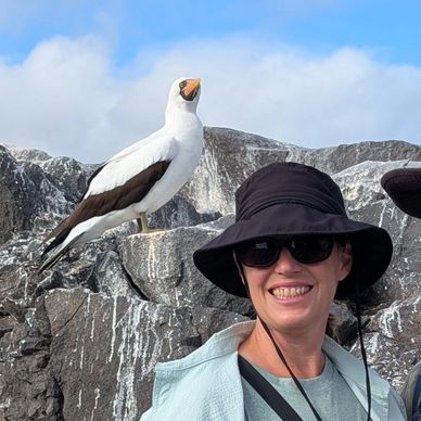 A smiling woman wearing sunglasses and a hat with a bird perched on rocks behind her.