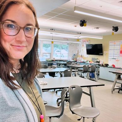 A woman smiling in an empty, well-lit classroom with modern chairs and desks.