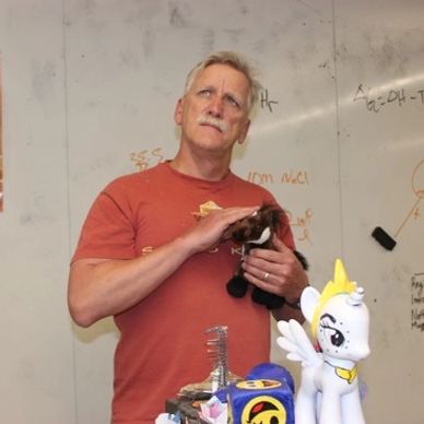 A man in a red shirt holding a small black cat in a classroom.