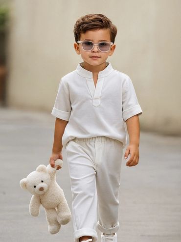 Young boy in white outfit holding a teddy bear and wearing sunglasses.