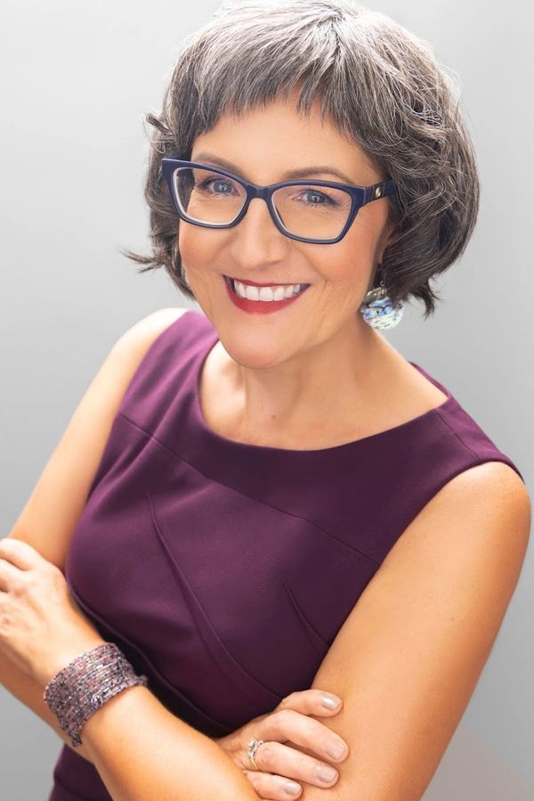 Smiling woman with glasses wearing a sleeveless purple dress and accessories.