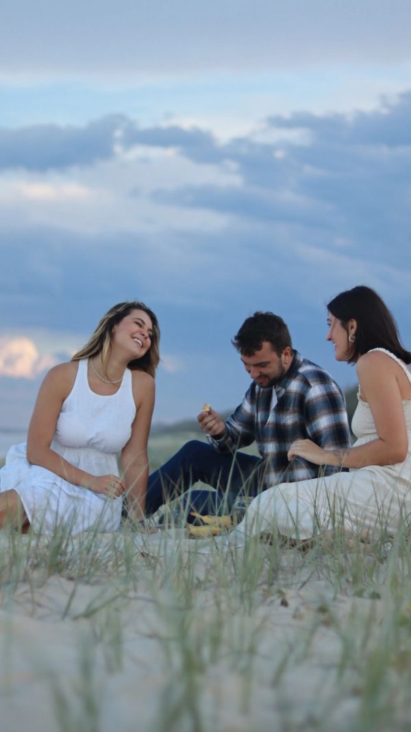 Three friends laughing and enjoying a picnic on a sandy beach under a cloudy sky.