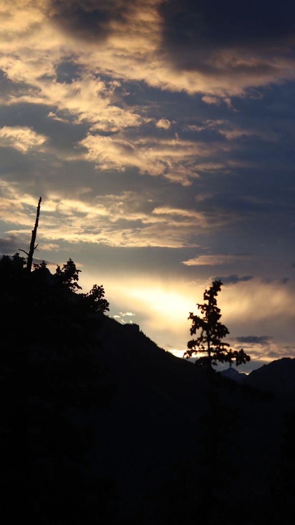 Sunset behind silhouetted mountains and trees with dramatic cloudy sky.