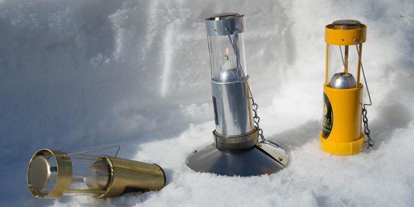 Three UCO Candle Lanterns in snow with Survival Stands attached. 2 standing, 1 tipped over.