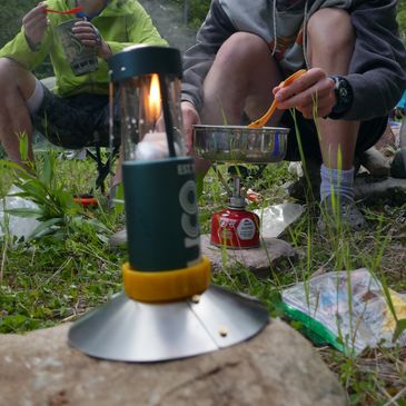 UCO Candle Lantern with Survival Stand attached sitting on rock outdoors with campers cooking 
