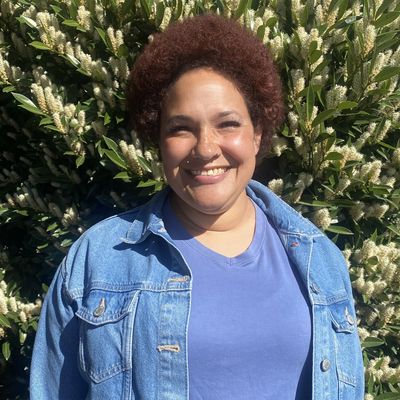 Smiling person with curly hair in a denim jacket outdoors.