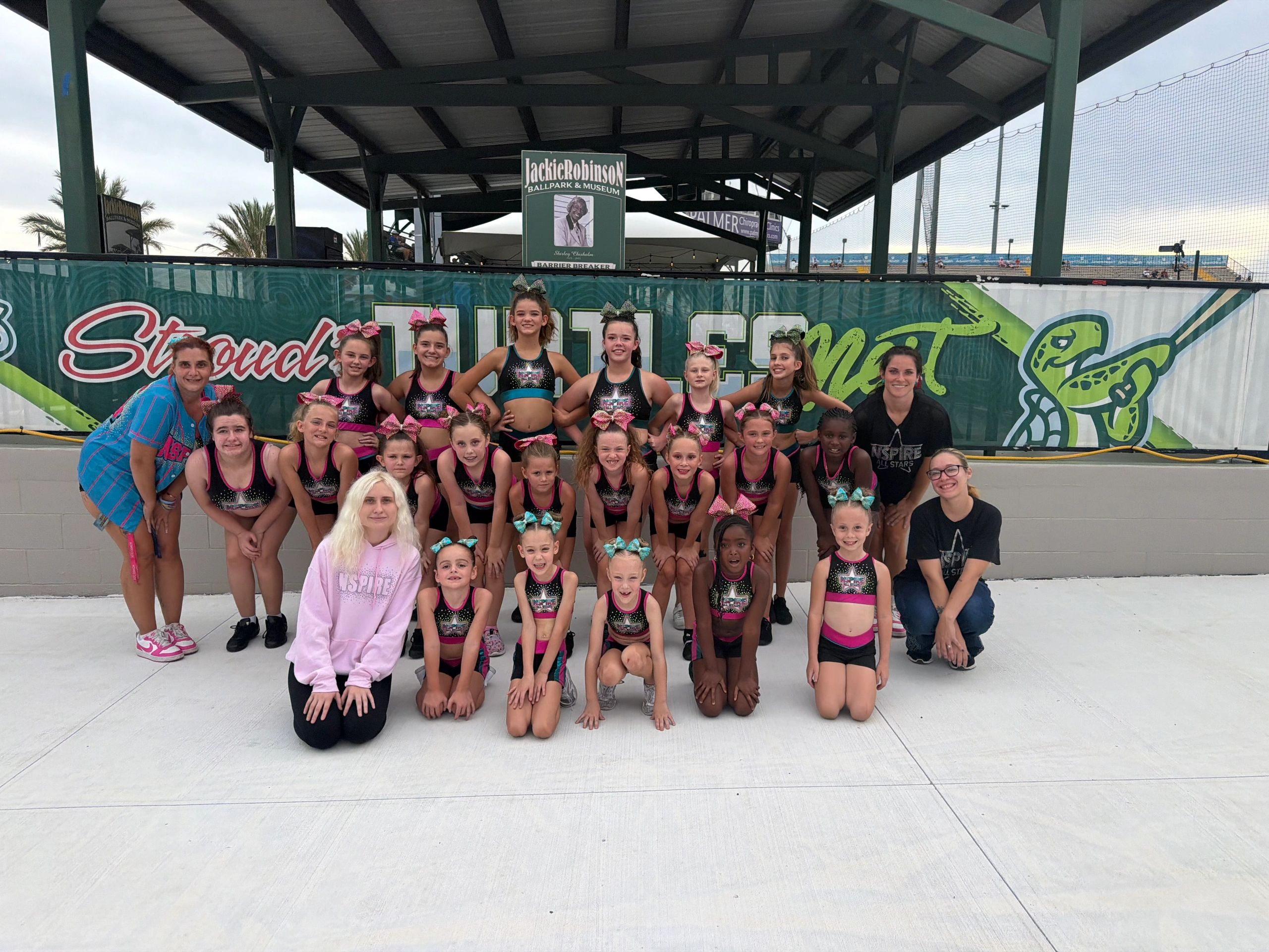 Group photo of young cheerleaders and coaches in front of a baseball field.