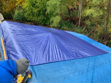 Person securing a blue tarp on a roof with a nail gun.