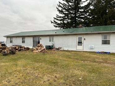 Single-story white house with green roof and firewood piles outside.