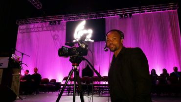 Man with headset operating a video camera on stage with purple lighting.