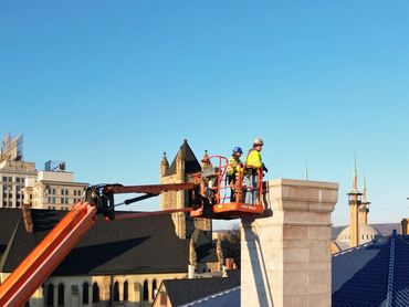 Workers on a lift inspecting a chimney on a blue-tiled roof under clear skies.