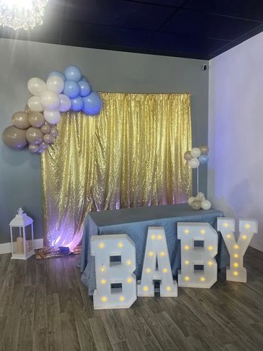 Gold sequin backdrop with a balloon garland and light up marquee "B-A-B-Y" letters for a baby shower.