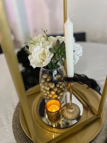 Elegant centerpiece with white flowers, gold accents, and a lit candle on a reflective tray.