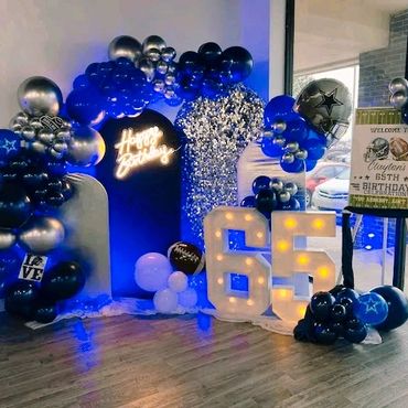 A 65th birthday party setup with blue, silver balloons, and illuminated numbers.