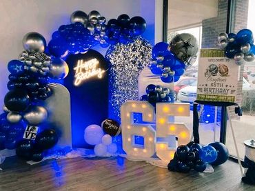 A 65th birthday party setup with blue, silver balloons, and illuminated numbers.