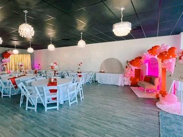 Decorated event space with white chairs, round tables, chandeliers, and balloon arrangements.