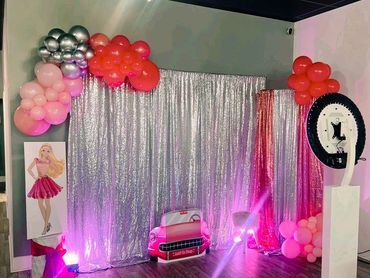 A shiny photo booth setup with pink balloons, silver backdrop, and a car cutout.