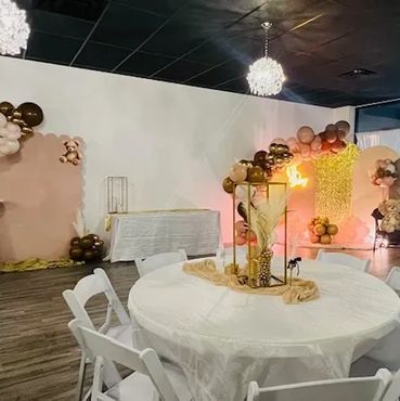 Elegant event space with white chairs, round table, and balloon decorations in warm tones.