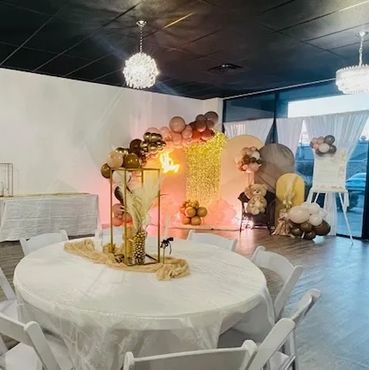 Elegant event space with white chairs, round table, and balloon decorations in warm tones.