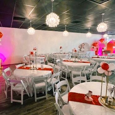 Elegant event setup with white tables, chairs, and red decorations under chandeliers.