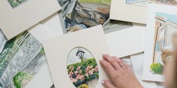 A hand arranging watercolor paintings featuring landscapes and flowers.