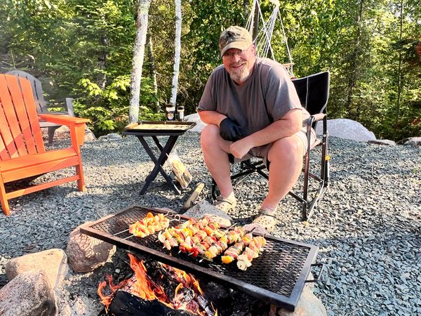 Man grilling skewers over an open campfire in a forest setting.