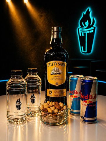 A bottle of Cutty Sark whisky with Red Bull cans and a bowl of nuts on a table.