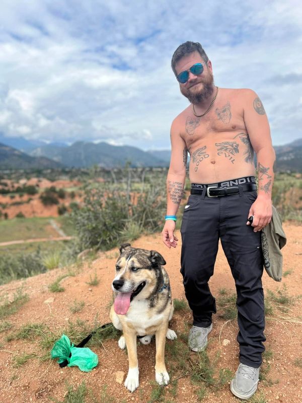Man with tattoos and sunglasses standing with a happy dog outdoors.