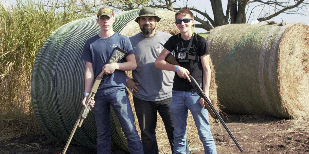 Three men posing outdoors with shotguns in front of large hay bales.