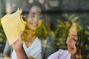 Two women cleaning a window with cloths, reflected greenery visible.