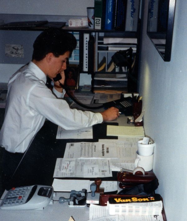A man in a white shirt working at a cluttered office desk while talking on the phone.
