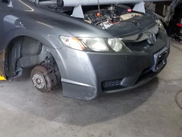 A gray Honda car with its hood open and front wheel removed in a garage.