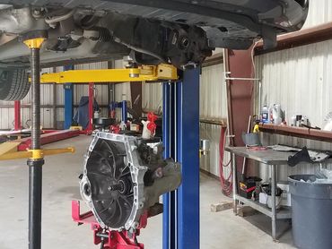 Car lifted in a garage with transmission removed and supported by a jack.