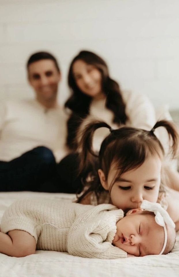Toddler kisses newborn baby on the cheek with parents blurred in background.