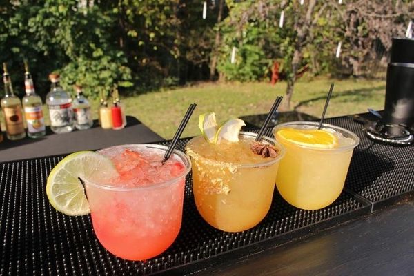 Three colorful cocktails with fruit garnishes on a bar mat outdoors.