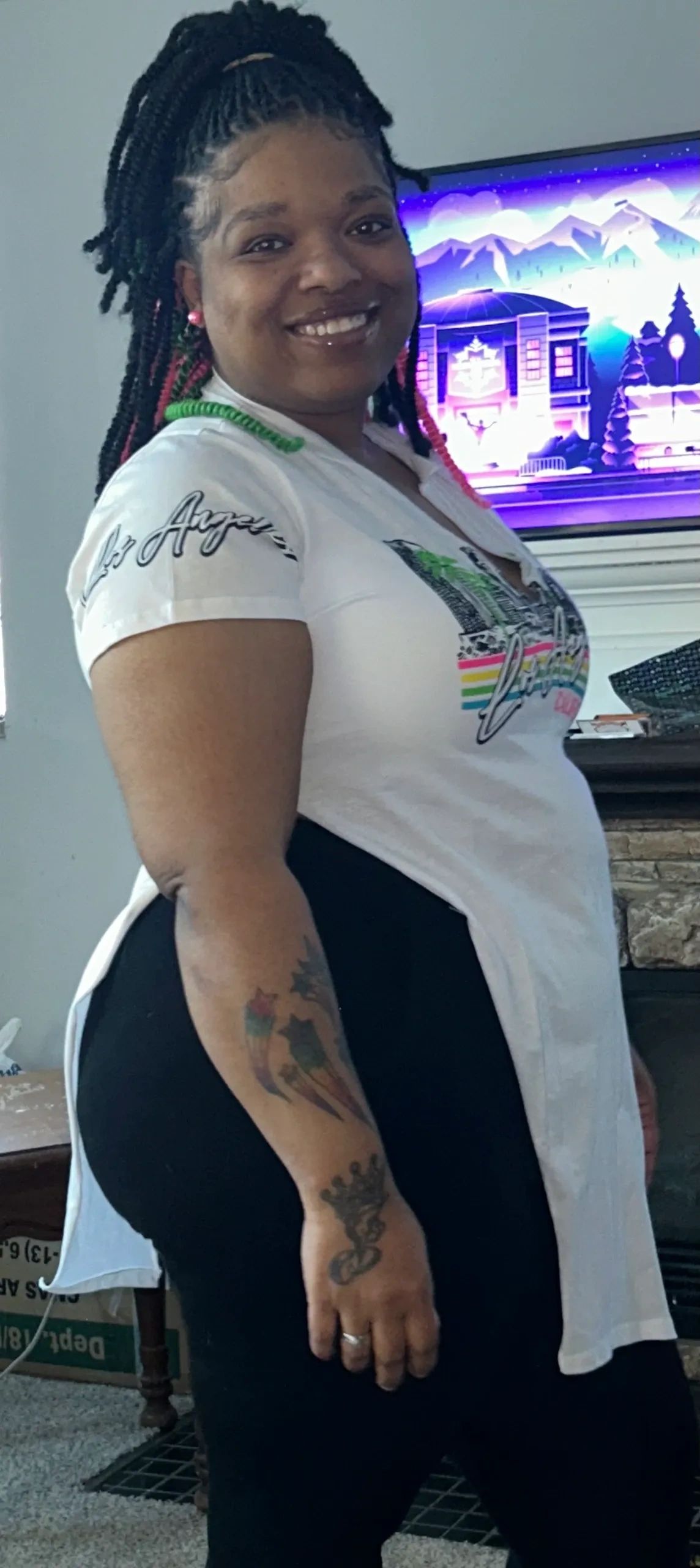 Smiling woman with braided hair and tattoos, wearing a white shirt and black pants indoors.