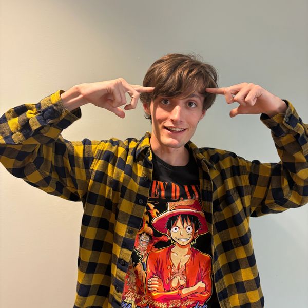 Young man pointing at his head, wearing a yellow plaid shirt and anime t-shirt.