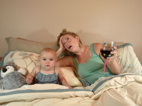 Tired mother in bed with baby and glass of wine, looking exhausted.