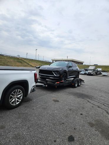 Black SUV loaded on a trailer attached to a white pickup truck in a parking lot.