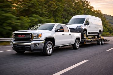White GMC pickup truck towing a white van on a trailer down the highway.