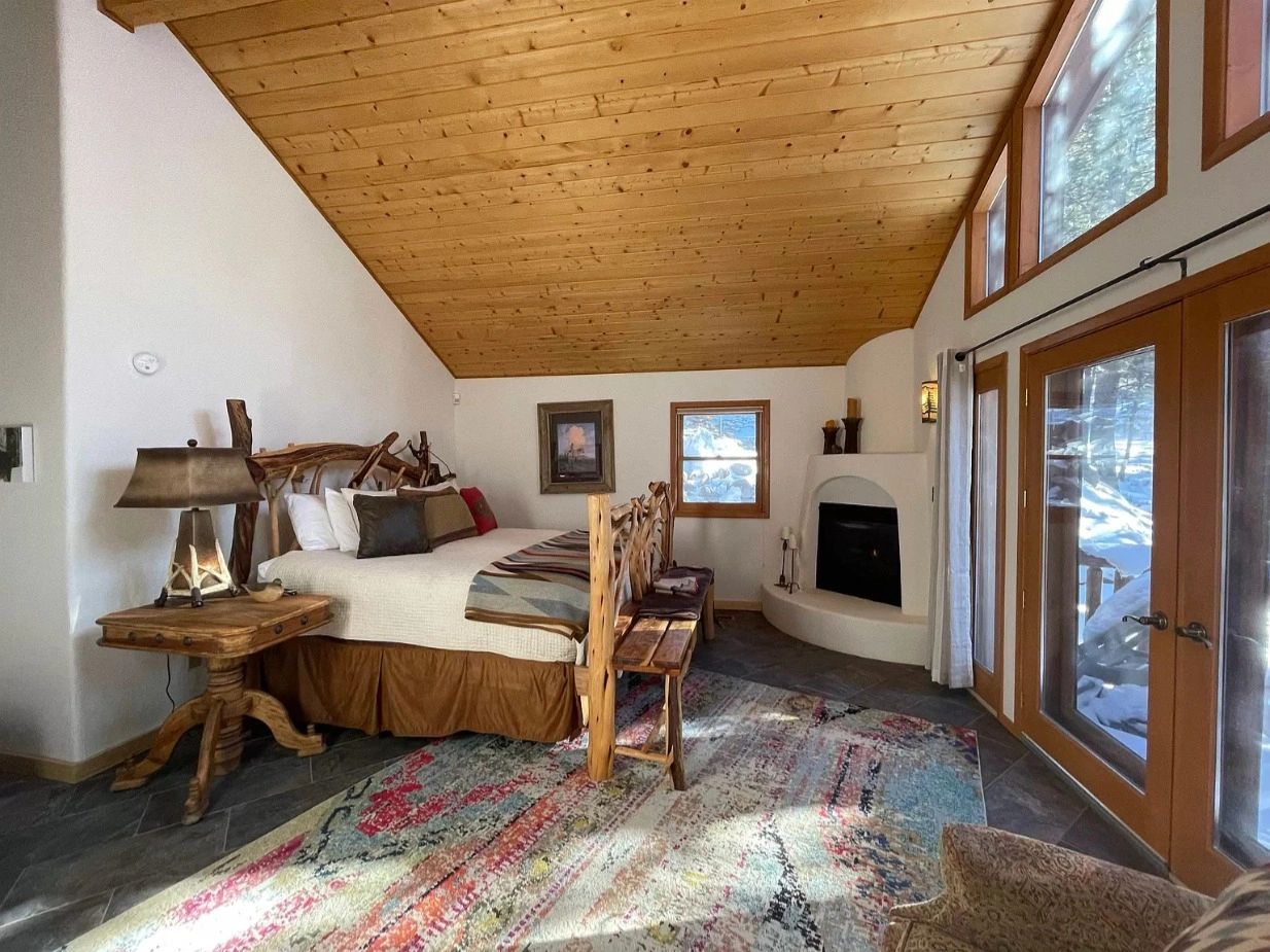 Rustic bedroom with wooden ceiling, fireplace, and large windows overlooking snow.