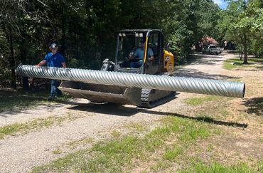 Two men handling a long corrugated metal pipe with a small tracked loader.