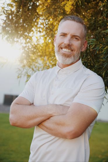 Smiling man with a white beard and polo shirt posing outdoors with arms crossed.