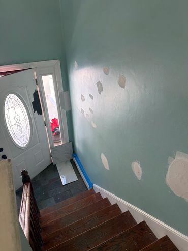Staircase with patched wall and open front door showing outside plants.
